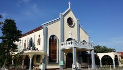 The external structure of the Carmelite Church shows white walls, red roof, and blue highlights on the pillars and edges. The roof is triangular with a cross on top-front-center. Under the cross is a flower-like circular glass window with eight petals. Another cross under the circular window is on the wall above a semicircular window that is partially covered by a balcony. The main entrance, a brown semi-oblong door, is under the balcony. There are also several similar doors on the sides as well as semi-oblong glass windows on both sides of the balcony.
