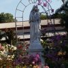 A white statue of Mary holding baby Jesus. They are surrounded by several flowers and plants in an open area. Steel arcs also form a frame around Mary. The red roof of Carmelite Church is visible in the background.