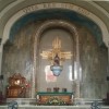 The sanctuary is mostly white, brown, and gold. The altar is color brown with green cloth on top. The sits and the pedestal are brown woods with gold-colored decoration. The statue at the center is that of Jesus sitting on a throne, with golden rays radiating from his back.