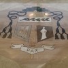 A bishop's coat of arm on the floor of Shrine of Mary, Queen of Peace. It has a pentagon-shaped shield at the center; It has twelves tassels, six on the left and six on the right of the shield. It has a cross on top of the shield and a galero hat above the cross. Below the shield is a ribbon strip with the word PAX, Latin for peace written on it. On the shield, there are figures: near the top is that of a plant branch with ten leaves and five pearl-like small spheres, on the left is what looks like a white silhouette of a bishop holding a book, and on the right is a large letter M.