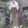 A monochrome statue of St. Francis of Assisi in one of the open air areas of EDSA Shrine. Here. St. Francis is holding a large clamshell, with two small birds on its side. The statues of the birds are positioned as if they are drinking water from inside the shell.