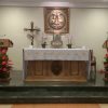The sanctuary of the Chapel of the Holy Family has two steps of black marble. At the center is a beige altar with dark brown highlights that look like four Greek columns and an oval protrusion with patterns at the center. It is draped with white cloth decorated with yellow crosses with rings (Celtic cross). It has two beige and brown stands with microphones: one on the left and one on the right of the altar. Each of these stands has a bouquet of red flowers in front, and what looks like a brown candle holder mounted on the wall at the back. A silver crucifix is also seen on the left (as seen from the nave), and the priest's chair is partly visible behind the altar on the right. An emboss mural of the Holy Family is mounted on the wall at the center behind the altar. The tabernacle is a silver box below the mural.