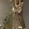 A statue of Mary is dressed in a white robe and long yellow-green hood with floral patterns. She is depicted as carrying baby Jesus in a white robe with her left hand, and a goldtone wand with her right hand. Both Jesus and Mary are wearing crowns proportional to their heads. Mary also has goldtone rays in front of her face and a halo with twenty-four stars attached on the back of her head.
