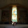 A tall and narrow stained glass shows Mary on clouds with three angels in front of her feet and yellow rays radiating from the top. Also seen in this picture is the loft at the back of the nave with what looks like a statue of resurrected Jesus attached to it. The paintings on the wall on the sides of the stained glass show various things but the wall is darker, so they are barely visible. These things are Mary talking to an angel; two hands holding a rosary; maps of France, Italy, and Spain; a wooden ship; priest giving a sermon, a gray church that looks like Manila Cathedral, and a group of cardinals with yellow mitres.