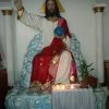 A large statue of Jesus depicts him sitting on blue and white clouds while holding a blue orb, possibly a representation of Earth, with his left hand, while his right hand extends diagonally upward. He is wearing white and red here with gold lining near the ends. A goldtone triangle is on the wall aligned to the head of Jesus. Two white pillars are on the sides of Jesus.