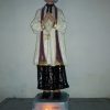 A statue of St. John Vianney shows him praying in Amen pose while looking up. He has long hair that reaches up to his neck. He is wearing a black internal cassock and on top of it, white surplice with goldtone lining at the end of sleeves. He is also wearing a violet stole over his shoulders. A candle with a live flame is in front of the statue.