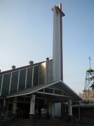 The external structure of Our Lady of La Paz church. It has triangular roof and triangular shade at the entrance. It's cross extends to height that seems like double the height of the main structure. A separate structure contains the bells.