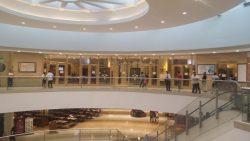 The outside of the Chapel of the Eucharistic Lord as seen from the other side of the round balcony. The face of the chapel is made of glass, through which the interior can be seen: the big LCDs inside are the most visible. The central door is between two glass panels with goldtone crosses on frames: one cross and frame on each panel. The words Chapel of the Eucharistic Lord is in metallic goldtone letters above the central entrance and glass panels with crosses.