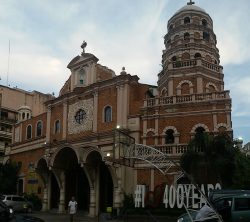 The Our Lady of Pillar Church has a pink and orange facade. It has three large semi-oblong doors in the main entrance and several semi-oblong windows framed with pink concrete arches. Above the main entrance is a small circular glass window made of nine glass panels. Above the circular window is a platform with what looks like a statue of Mary. On the highest part of the front wall above the statue, is a decoration that looks like the blessed sacrament with a small cross on top. On the right of the church (when viewed in front from outside) is a large bell tower with similar orange and pink design and has several semi-oblong windows surrounding it. Its height exceeds that of the main church structure and has a small cross on top. There is also a smaller white bell tower on the left. Its height is lower than the main church structure.