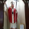 A statue of Pope John Paul II is inside a small shelf framed with maroon wood. The figure depicts the pope as raising his right hand while holding a staff with a crucifix with his left hand. He is wearing a white alb, red chasuble, and white stole decorated with two small black crosses, a black pentagon shield, and what looks like goldtone keys forming X. His brown boots are visible under the alb. A yellow picture frame with a text of Prayer to St. John Paul II is leaning on the wall on his left.