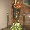 Statue of Mary carrying child Jesus and both wearing a crown. Mary is dressed in green and orange while Jesus in red and brown. There are flowers in front of Mary's foot and candle holders on her sides.