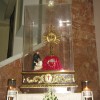 The monstrance inside a glass container at Our Lady of La Paz Parish: it is gold or bronze in color and shaped like sun disc with emanating rays. There is a cross on top of it, and it is mounted on a pedestal covered with red cloth. There is a statue of what appears to be a saint helping another person or maybe a depiction of the good Samaritan.