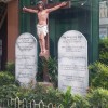 A plant box outside OLPH Parish with two white tablet stones containing the ten commandments on the left and the right of a crucifix. The ten commandments are arranged in an unusual manner, with the first five commandments on one stone and the last five on the other stone, unlike the usual three and seven. The commandments are written in Filipino, with English translations underneath each of them in a smaller font and enclosed in parentheses. The crucifix is of different shades of brown except for Jesus's white underwear, red blood from his wounds, and the white INRI signage near the top. The tablets and the crucifix are surrounded by plants, and the plant box is enclosed by white iron railings.
