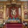 The altar of Our Lady of Perpetual Help Parish in Cubao is primarily that of gold and red color. At the center is a brown cross on red wall background, with rays emanating from its center. In front of it is a statue of Jesus, but this is not a crucifix because Jesus is fully clothed and his feet are not nailed together; looks like a depiction of his Ascension rather than Crucifixion. There are two angels, one on the left and one on the right of Jesus' feet, and two additional statues of angels on the bottom left and bottom right of the cross. In front of Jesus is a gold-colored altar with green cloth on top. On the sides are two Sun-like decorations; one with embossed figures of two angels facing each other below it, and the other has an icon of Mary carrying baby Jesus below it. The center area of the sanctuary in front of the altar as a red carpet. There are also two podiums for lectors and commentators; one on the lower platform of the sanctuary, while the other in front on the same level as the nave.