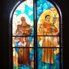 A semi-oblong stained glass pane as seen from inside the San Antonio De Padua Parish is divided vertically into two sections by a steel frame. The left half depicts St. Anthony of Padua holding baby Jesus on the shoulder with his right hand. He is wearing a brown sackcloth robe with a rope as a belt. Jesus is sitting on a mass of cloud hovering in front of St. Anthony. Part of the cloud also holds a red book. St. Anthony has a rosary attached to the left part of his belt. Jesus is wearing a white robe. The right half of the stained glass also depicts St. Anthony, with the same clothing as in the left half. Here, he is standing while holding a twig with white lilies.