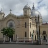 Shown here is the front of San Diego De Alcala Parish, with a view slightly angled so that its right side is partly visible. The facade of the church is mainly mocha in color, with red on top of the triangular roof and gray on the spherical domes. There are three spherical domes, two smaller ones in the front: one serves as a clock tower and the other as a bell tower. Between them is a triangular part of the roof with a cross on top. The larger and taller dome at the back is above the sanctuary inside the church; it has a cross on top. The church has several small windows. It is surrounded by a white fence made of iron, with statues of the apostles on every two pillars holding the fence.