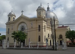 Shown here is the front of San Diego De Alcala Parish, with a view slightly angled so that its right side is partly visible. The facade of the church is mainly mocha in color, with red on top of the triangular roof and gray on the spherical domes. There are three spherical domes, two smaller ones in the front: one serves as a clock tower and the other as a bell tower. Between them is a triangular part of the roof with a cross on top. The larger and taller dome at the back is above the sanctuary inside the church; it has a cross on top. The church has several small windows. It is surrounded by a white fence made of iron, with statues of the apostles on every two pillars holding the fence.