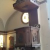 The pulpit of San Diego Parish is made of brown wood and has murals on the side. It also has a white microphone or speaker mounted on its balcony and a ceiling-like structure above. The ceiling-like structure has a dove mural underneath. The underside of the pulpit has a design that looks like a curly Roman pillar.