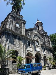 The front of San Sebastian Cathedral as seen from the corner of its yard. It has gray facade and has two towers on the sides. The curvature of the towers is due to vertical panorama shot; they are actually straight. It has several semi-oblong windows and also a semi-oblong main entrance at the center and ground level. Above the main entrance is a circular cavity with what looks like a white statue of Jesus inside. The central structure between the bell towers has a triangular roof with a cross on top. The bell towers also have crosses on top and circular windows below the bell structures.