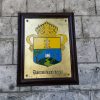 A goldtone metal plate shows the seal of the Diocese of Bacolod; an escutcheon divided vertically into two halves; yellow and blue. The yellow on top has a white cross with two brown arrows penetrating it at its foot. The blue half has a hill with a church on top and trees on the side. The escutcheon has a crown on top.