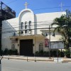 This is the frontage of Santisima Trinidad Parish along Estrada St. It has white facade with semi-oblong arc in the middle. A cross is on top of the semi-oblong and below it is the brown rectangular main entrance. The ground on both sides of the entrance is decorated with potted plants.