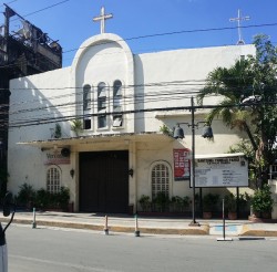 This is the frontage of Santisima Trinidad Parish along Estrada St. It has white facade with semi-oblong arc in the middle. A cross is on top of the semi-oblong and below it is the brown rectangular main entrance. The ground on both sides of the entrance is decorated with potted plants.