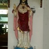 A statue of Jesus with sacred heart. Jesus here wears a white robe with red layers over it. His right hand (left of view) forms a peace sign, while his left hand is pointing to his sacred heart, which has golden rays emanating from it.