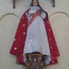 A statue of Sto. Niño. He has long brown hair and wears a crown. His clothes are made of a white robe with a red cape and a red sash. He is holding a brown rod and brown orb with cross on top.