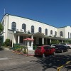 Shown here is the side and part of the front of Santuario De San Jose as seen from Buffalo St. The structure of the church is long and painted in white with a green roof. The upper part of the side has a row of several semi-oblong stained glass windows showing different events from the Bible. The lower part has large wooden gates, ramps for wheelchairs, and a small house-like structure with glass on sides and a pink roof. There are also several cars parked on the side of the church in this picture.