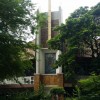 The structure of St. Francis Church as seen from across the street. It is blocked by several trees and other plants on the island dividing the street and those that are in front of it. The church is elevated; there are doors on the ground level, but the actual church is at the second story of the building accessible through ramps on both sides. The church has white frontage and brown roof as well as yellowish bricks at the central structure above and below the glass pane of St. Francis, which is at the center of the frontage. A yellow cross is on top and upper front of the central structure.
