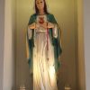A statue of Mary showing her Immaculate Heart inside the Chapel of St. Padre Pio in Shangri La EDSA. She is wearing a white robe, white hood, and cyan cape. She is holding a red rosary with left hand and a white rosary with right hand; the rosaries are hanging freely on the ends with crosses. Also visible on the sides of her feet are two cylindrical glass candle holders with candles inside and her portraits showing through the transparent glass.