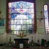 The sanctuary of San Isidro Labrador Parish is dominated by a yellow-green wall and below it a long white partition. The center of the yellow-green wall has stained glass images of God the Father in the form of an old bearded man with stretched arms. In front of the Father is the Holy Spirit in dove form. Below them are panes of clear glasses through which the crucifix outside the physical structure of the parish is visible, surrounded by plants. At the center of the partition is the tabernacle flanked by yellow flowers. In front of tabernacle is the altar with a green cloth cover. There are doors on the left and right of the partition that seems to be access ways to whatever is behind the partition, maybe the sacristy. There is also a small wooden podium on the right and a larger podium on the left (if facing the sanctuary) for the speakers: priests, lector, or commentators.