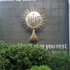 A bronze ornament that looks like the Sun with letters IHS on the sun disc mounted on a gray wall. It also has a cup-like stand although it is mounted on the wall outside the San Isidro Labrador Parish's structure. It is surrounded by the text 'Come to me...' (above) and '...and I will give you rest' (below) from Matthew 11:28.
