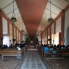 The interior of the St. John the Baptist Parish is mostly brown and white on the roof, walls, and floor. Seen here is the corridor leading to the sanctuary. Several rows of sits for the laity are grouped to left and right sides of the corridor. Also visible here are six chandeliers hanging on the ceiling.