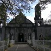 The frontage of St. John the Baptist Parish as seen from J. P. Rizal St. in Calamba, Laguna. A long corridor surrounded by white grills and plants separate the church's main entrance from its gate. The church is gray with visible bricks and towers on the left and right of the church's main structure.