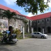 The side of the external structure of St. John the Baptist Parish, showing several visible gray bricks, wooden semi-oblong doors, and red roofs.