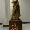 A goldtone statue of Joseph stands on a brown wooden pedestal in the sanctuary of Our Lady of the Most Holy Rosary Chapel. Joseph is holding a staff with three flowers at the end. There are white flowers and red and green leaves in front of the wooden pedestal.