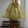 A goldtone statue of Mary carrying baby Jesus stands on a brown wooden pedestal in the sanctuary of Our Lady of the Most Holy Rosary Chapel. Mary has a halo on the back of her head that radiates outward and end with stars, and also a separate crown on top of her head. The baby Jesus statue is also goldtone and wears a smaller crown. There are white flowers and red and green leaves in front of the wooden pedestal.