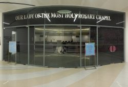 The exterior of Our Lady of the Most Holy Rosary is made of glass walls and doors. The words Our Lady of the Most Holy Rosary Chapel is written prominently above the window. The nave and the sanctuary are visible through the glasses. This picture is a panoramic version; the glass walls are actually flat in shape and not curved.