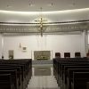 The sanctuary and parts of the nave are seen in this picture. As seen from the nave, the altar is at the center; it is a gray stone structure with candle stands on both sides and yellow and white flowers in front. A lectern with similar color as the altar is on the left. The tabernacle is a small goldtone box attached to the wall between the altar and the lectern. On the left of the lectern is a goldtone stature of Mary carrying baby Jesus. There are three seats on the right of the altar, and on the right of them is a goldtone statue of Joseph. A brown wooden podium with microphone, also used by lectors and commentators, is standing in front of St. Joseph's statue. A goldtone crucifix is hanging on the ceiling above the altar.