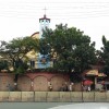 The Sto. Rosario Parish as seen from the opposite side of Ortigas Avenue. The church is mostly yellow with maroon roof and blue bell tower at the center. The church structure is covered by a salmon pink-colored wall with a large gate on the right side. Trees grow in front of the church, and their thick bush further covers the church building.