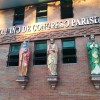 Close-up of the external red wall of Sto. Niño De Congreso, showing the letters STO. NINO DE CONGRESO PARISH on metallic letters embedded on the wall just below the upper rows of windows. Below them are the lower rows of windows: four windows are seen in this picture. There are statues of apostles St. James the Greater, St. Philip, and St. Thomas, on the wall between the windows: one apostle between two adjacent windows. St. James is wearing red and orange, has graying hair, and holding a sword. St. Philip is wearing red and beige and embracing what looks like a plank of wood. St. Thomas is wearing green and yellow, in amen pose while embracing what looks like a fishing spear.
