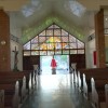 View of the main entrance of Sto. Niño De Congreso as seen from the inside. The main entrance has a high triangular ceiling with loft visible on either side. It is mostly white but with two red-orange pillars where the lofts end and the high ceiling starts. The main doors are made of wood, and above them are glass panels from where the outside is visible. Just outside this glass panel is a glass pane of similar shape depicting what looks like angels in the clouds and yellow rays of light emanating from the top center. Each of the red-orange pillars has a small goldtone cross. A crucifix is also visible on the left (if you are viewing from the nave).