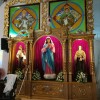 A gold-tone shelf with three panels contains statues and flowers on their feet. The one at the middle is the largest depicting Mary in pink robe, blue cloak, and white hood. The ones on the side are smaller. On the left is Jesus holding a lamb with both arms while tucking a brown staff on his elbow. I do not know who is this saint on the right: she is wearing a brown robe and cape and holding what looks like a monstrance. Above the shelf are portraits of St. Luke and St. John. St. Luke is holding a scroll with both hands, and a winged bull is partly visible on his left. St. John is holding a scroll with his right hand while letting its other end dangle; a while eagle is partly visible on his left.
