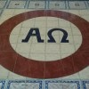 The characters Alpha and Omega in black surrounded by a brown circle and on a white background as part of the decorative tiles on the floor of St. Jude Thaddeus Parish.